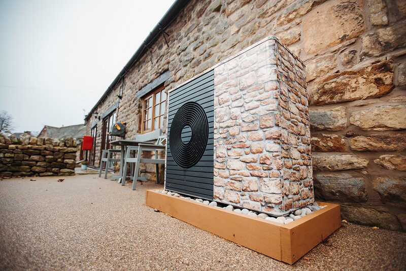 An air source heat pump unit with a stone-effect cover installed outside a stone cottage on a wooden plinth