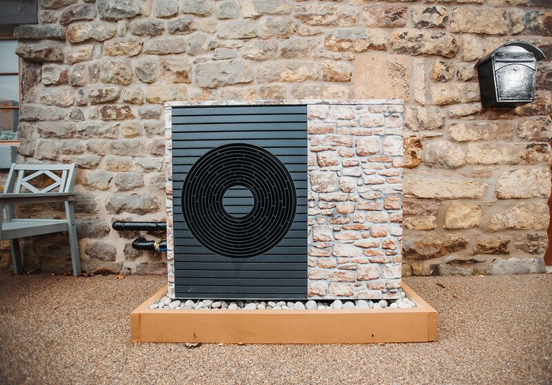 An air source heat pump unit with a decorative stone-effect cover installed outside a stone cottage on a gravel base