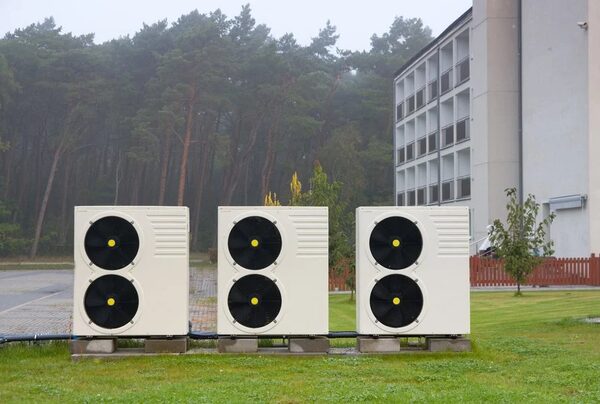 Three large cream air source heat pump units with black fans installed on grass outside a building