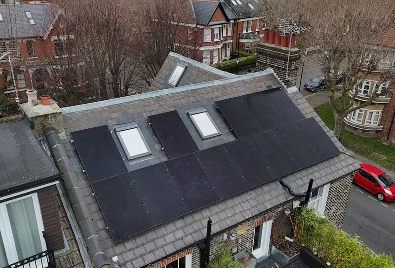 Black solar panels installed on the grey slate roof of a UK house, seen from above with neighbouring houses and a red car in the street below
