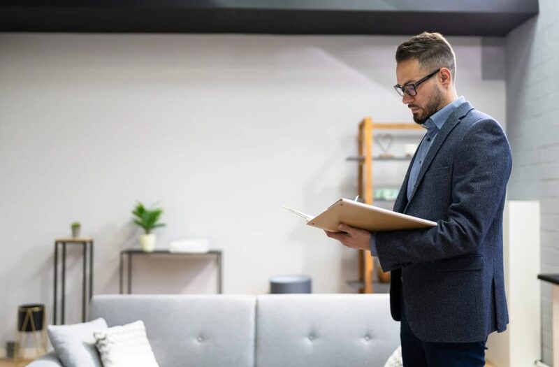 A man in a suit standing in a modern living room reading notes on a clipboard, as if conducting a survey or assessment