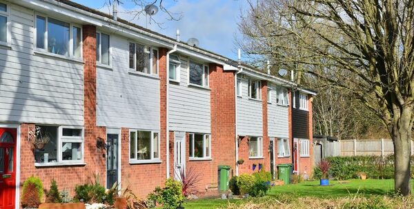 A row of brick and white-clad terraced social houses with satellite dishes, wheelie bins and front gardens on a sunny day