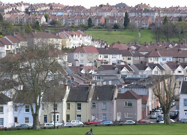 A view over rows of terraced and semi-detached houses on hilly streets in a UK suburb with parked cars and a park in the foreground