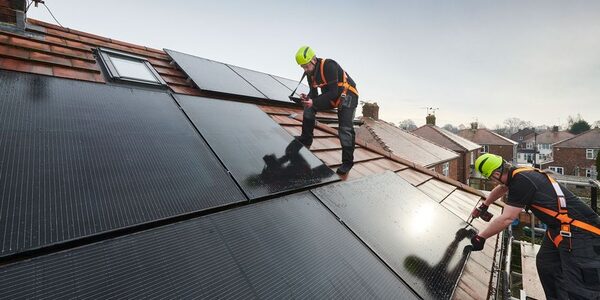 Two installers in safety harnesses fitting solar panels on the tiled roof of a UK house