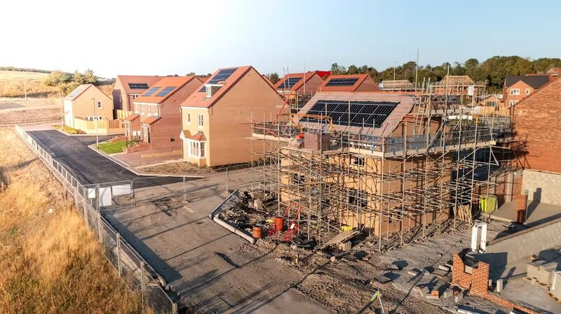 A new-build housing estate under construction with solar panels already fitted on several brick house roofs and scaffolding around unfinished homes