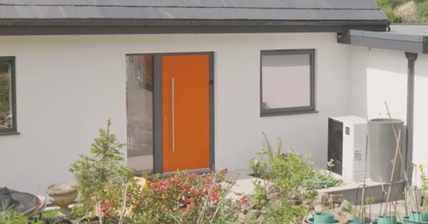 An air source heat pump unit installed outside a modern white-rendered house with an orange front door and garden plants