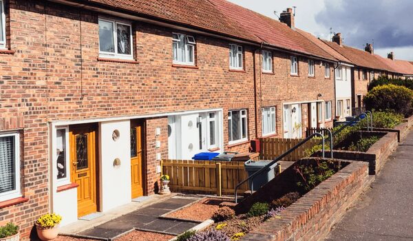 A row of brick terraced houses with front doors, gardens and wheelie bins on a residential street