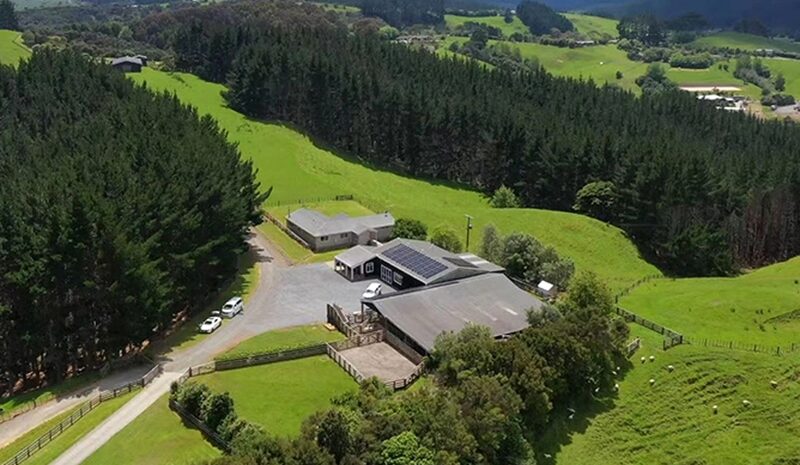 Aerial view of a rural farmhouse property with solar panels on the roof, surrounded by green fields and forest