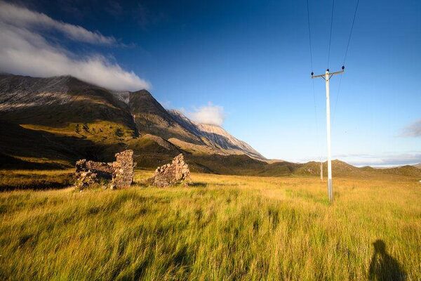 A wooden electricity distribution pole with power lines running through a grassy Highland landscape with ruined stone croft buildings and mountains behind