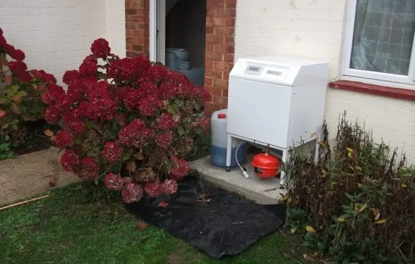 A white ground source heat pump unit installed outside a house next to a hydrangea bush, with an expansion vessel and a fluid container beneath it