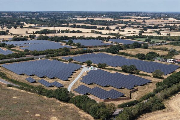 An aerial view of a large solar farm with rows of panels surrounded by countryside fields and trees
