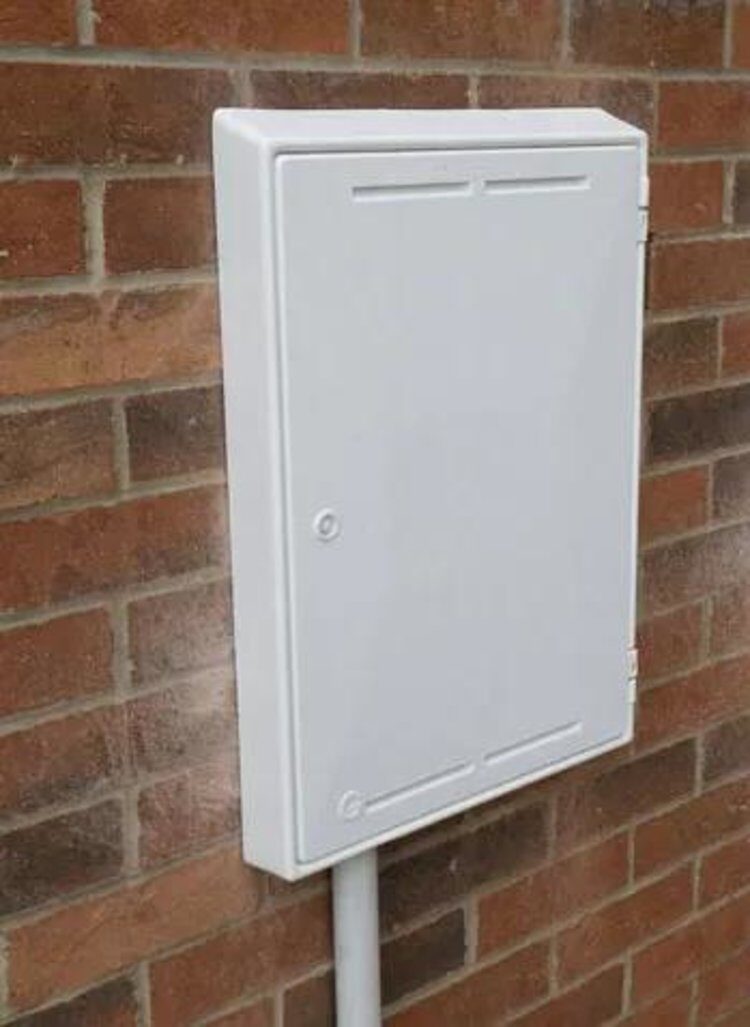 A white external electricity meter box mounted on a brick house wall with a conduit below