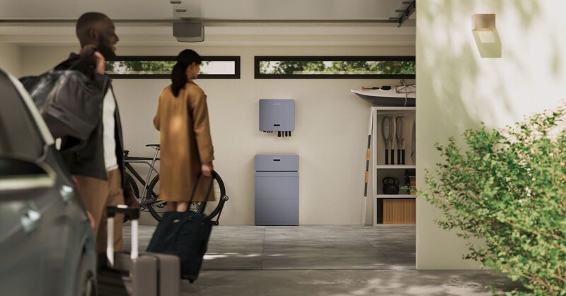 A wall-mounted home battery inverter and stacked battery unit installed in a garage, with a couple walking past with luggage