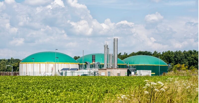 A biogas plant with three large green domed digesters and chimneys behind a green crop field under a cloudy sky