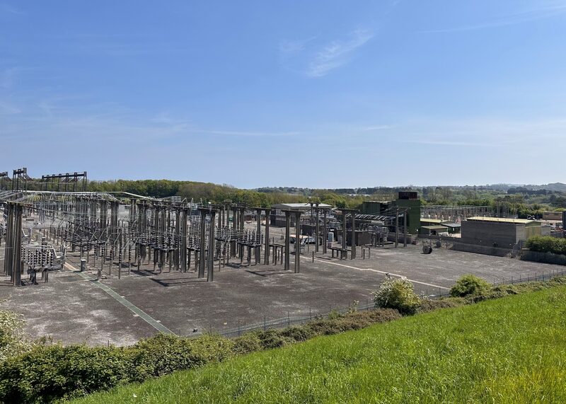 An outdoor electricity substation with pylons, switchgear and buildings seen from a grassy slope under a blue sky