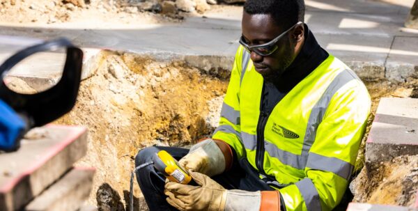 A UK Power Networks engineer in a high-visibility jacket and safety glasses working in an excavation trench with a cable detection tool