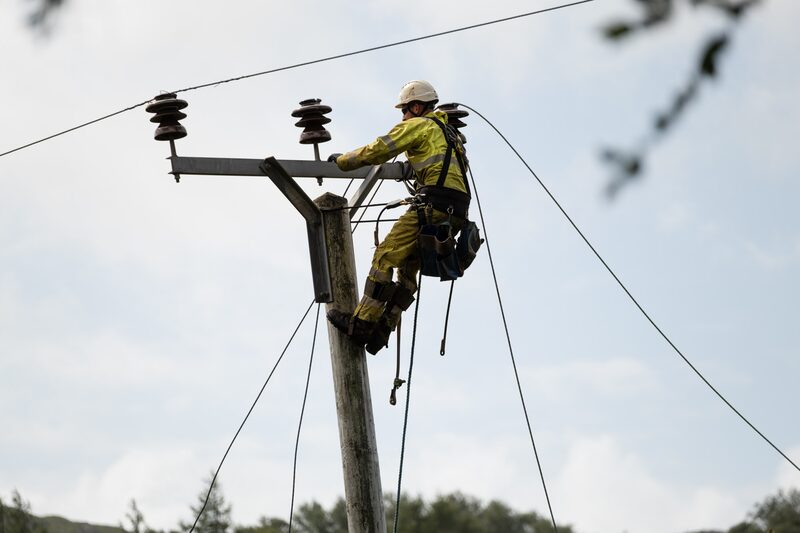 A linesman in high-visibility safety gear and helmet climbing a wooden electricity pole to repair overhead power lines