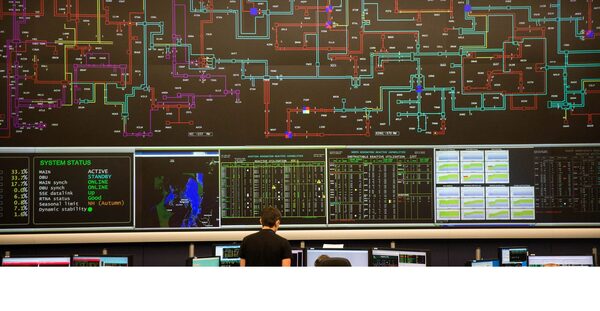 An electricity grid control room with a large wall of system status screens and schematic diagrams, with an operator seated at a desk below