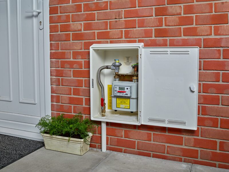 A gas meter installed in an open white meter box on the exterior red brick wall of a house beside a front door