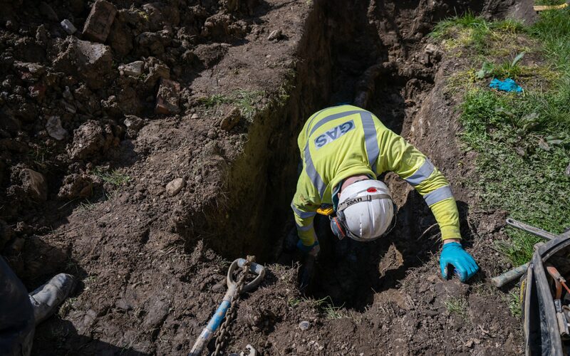A gas engineer in a high-visibility GAS jacket and white hard hat working in a dug trench laying gas pipework