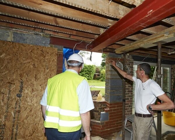A surveyor in a hi-vis vest and hard hat inspecting exposed floor joists and a steel beam with a builder at a house renovation site