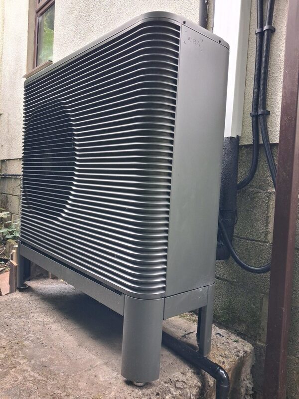 An Aira air source heat pump outdoor unit installed on a concrete base against the exterior wall of a house