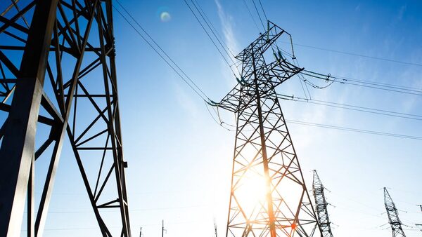 High-voltage electricity pylons and transmission lines against a bright blue sky with the sun shining through