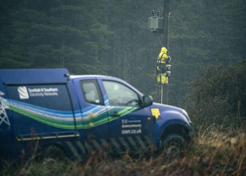 A Scottish and Southern Electricity Networks engineer in high-visibility gear works on a wooden power pole with a transformer while a branded SSEN utility pickup is parked in the foreground