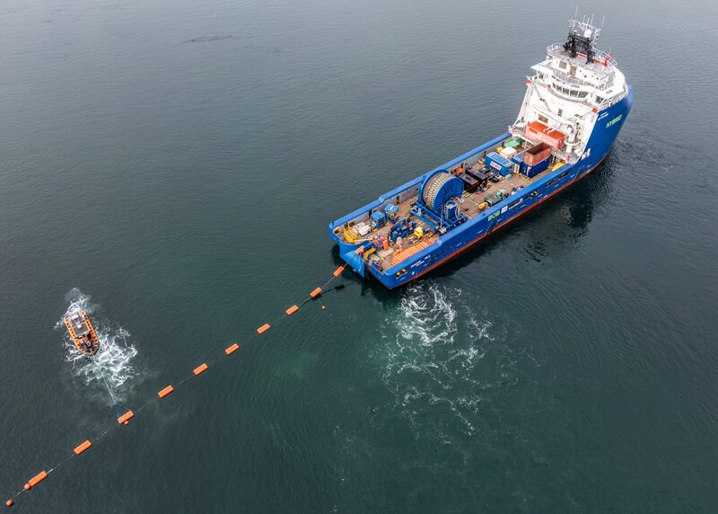 A blue cable-laying vessel laying a subsea electricity cable marked by orange buoys, with a small workboat alongside