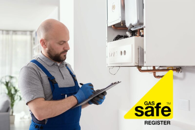 A Gas Safe registered engineer in blue overalls inspecting a wall-mounted boiler while taking notes on a clipboard