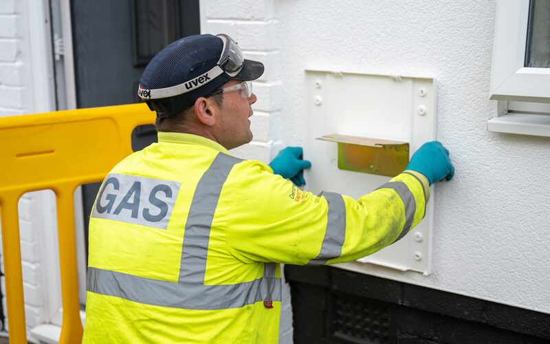 A gas engineer in a high-visibility GAS jacket and safety glasses working on a white gas meter box on an outside house wall