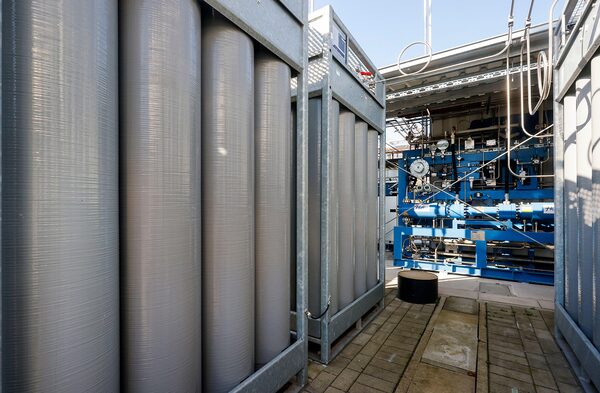Rows of large grey cylindrical gas storage tanks beside blue industrial pipework and valves at an outdoor hydrogen or gas processing facility