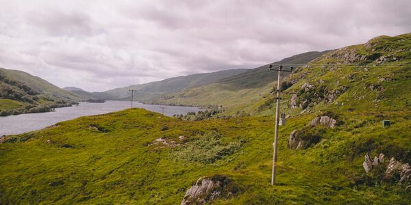 A wooden electricity distribution pole carrying overhead power lines across a green Highland landscape with a loch and hills under a cloudy sky