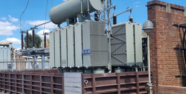 A large grey SGB electricity transformer at a substation, surrounded by flood-defence barriers next to a brick wall
