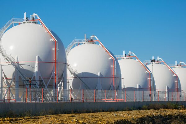 A row of large white spherical LNG storage tanks behind a fence under a blue sky