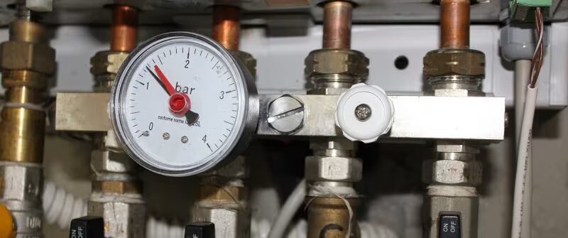 A close-up of a boiler pressure gauge reading in bar, mounted among brass valves and copper pipework on a heating manifold