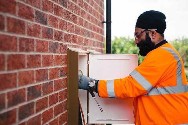 A gas engineer in a high-visibility orange jacket and turban working on a wall-mounted gas meter box on a brick house