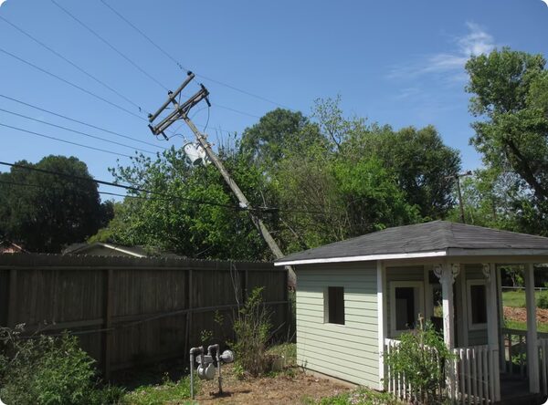 A wooden electricity pole leaning at an angle with power lines and a transformer, behind a garden fence and a small green shed