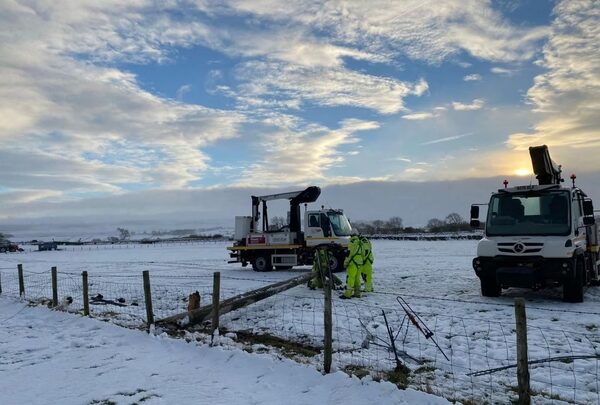 Utility workers in high-visibility clothing repairing a fallen power line in a snowy field beside two cherry-picker trucks after Storm Arwen