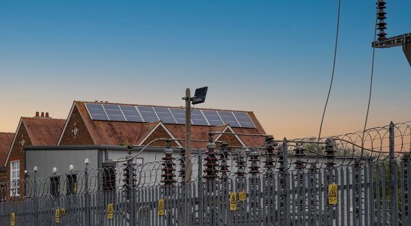 An electricity substation with barbed-wire fencing and insulators in front of a brick building with rooftop solar panels at dusk