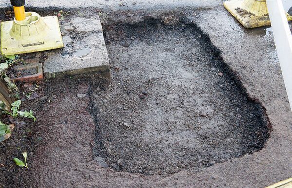 A dug-up patch of tarmac on a road or pavement, an excavation hole for gas works, with yellow jack leg supports at the edges