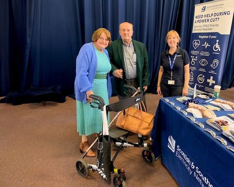 An SSEN staff member with an elderly couple and a mobility walker at a community event stand promoting the free Priority Services Register