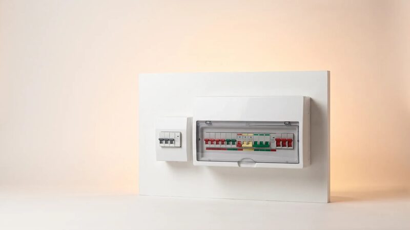 A white consumer unit (fuse box) with circuit breakers and a small switch panel mounted on a plain wall