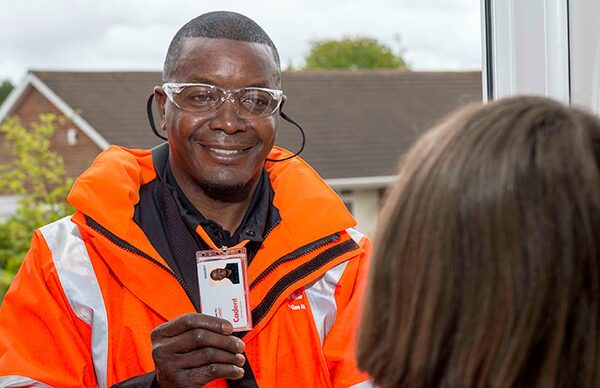 A Cadent gas engineer in an orange hi-vis jacket and safety glasses showing his ID badge to a customer at the door