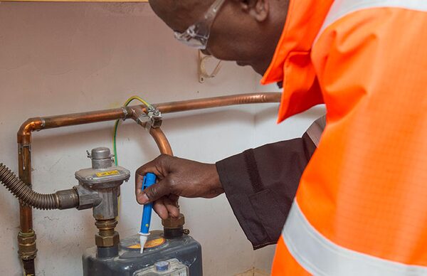 An engineer in a high-visibility orange jacket and safety glasses using a gas detector pen to check for leaks around a domestic gas meter and copper pipework