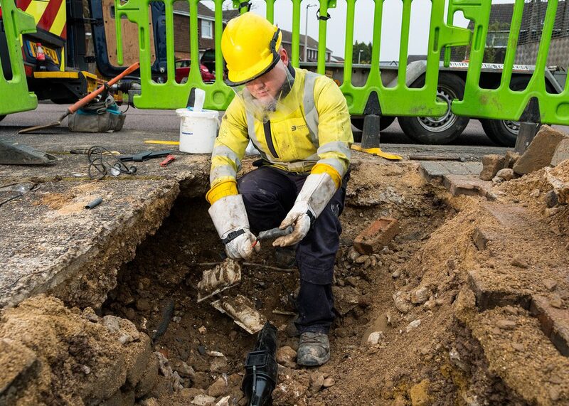 A utility worker in hi-vis clothing and yellow hard hat working in an excavated trench on underground power cables in a residential street