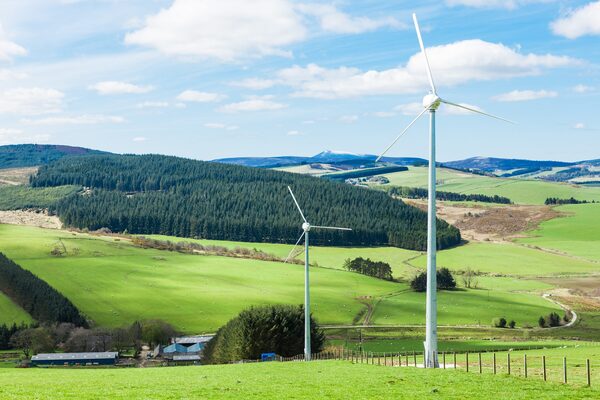 Two white wind turbines standing in green rolling countryside with hills, forest and a farm in the background