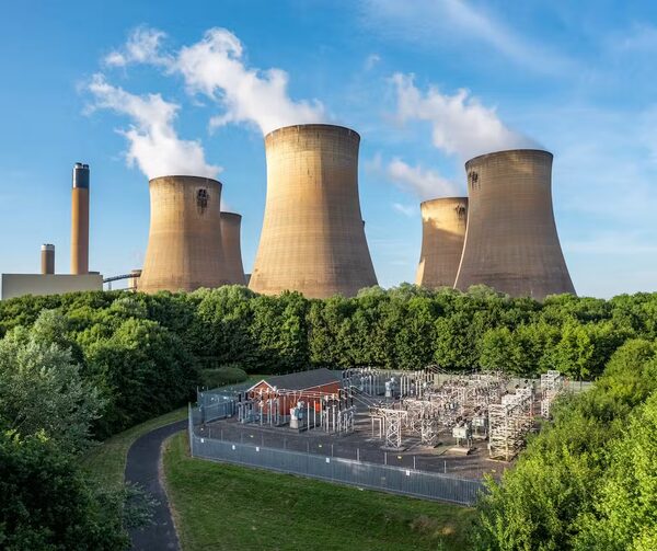 A power station with large cooling towers emitting steam, a chimney, and an electrical substation in the foreground behind trees