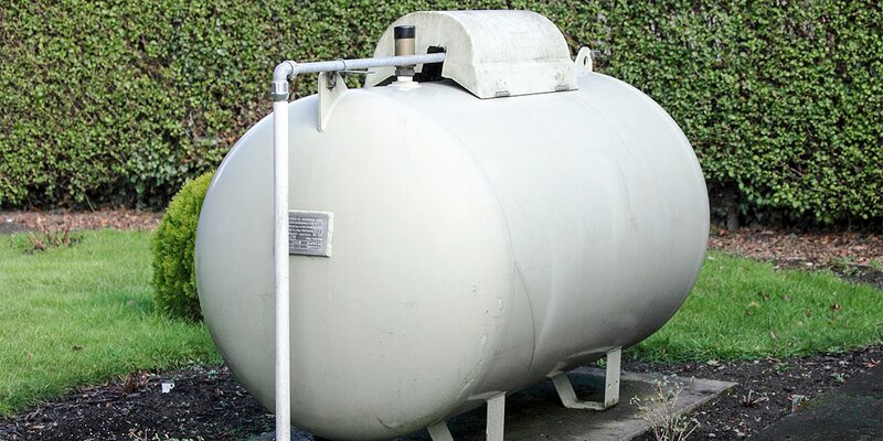 A large white cylindrical LPG storage tank on legs in a garden with a pipe and plaque, set against a hedge.