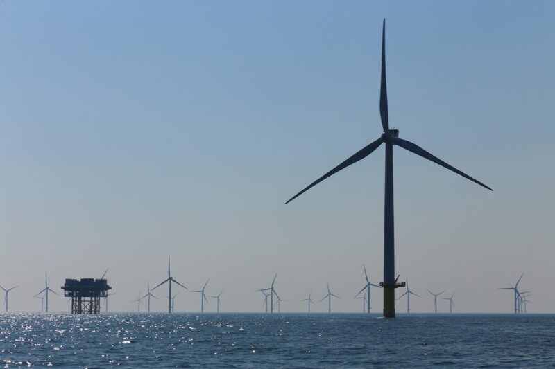 An offshore wind farm with a large turbine in the foreground and many smaller turbines and a substation on the sea horizon
